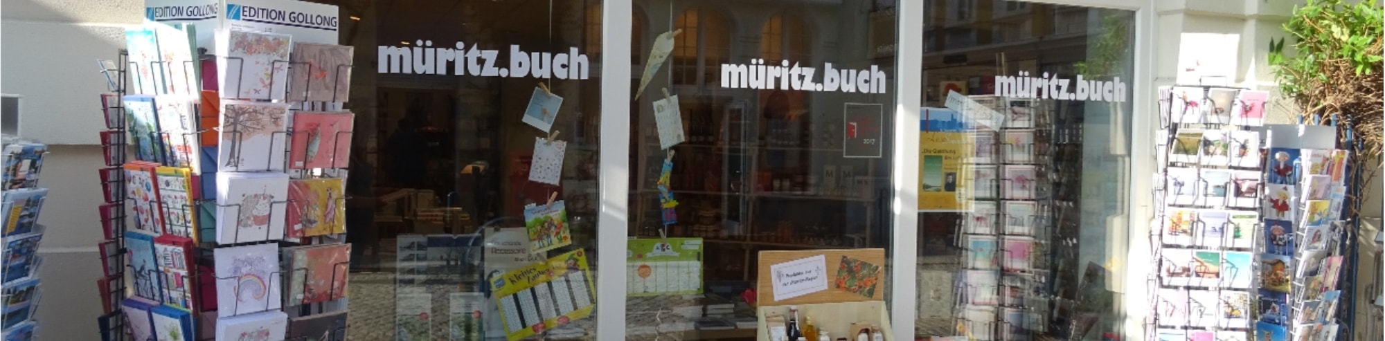 Foto des Schaufensters der Buchhandlung von außen, zu sehen außerdem Postkartenständer